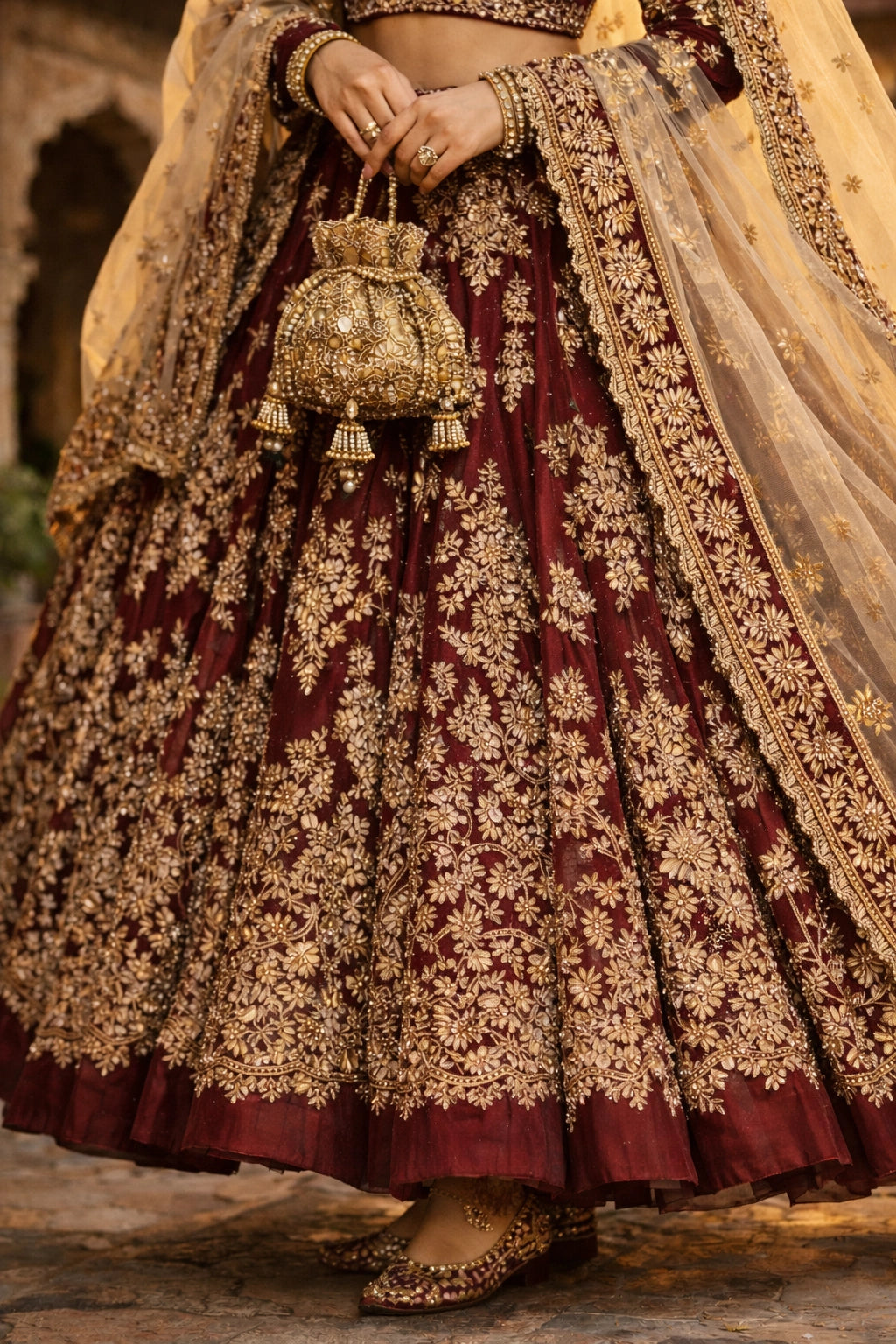Bride wearing deep maroon bridal lehenga with gold embroidery and sheer dupatta in heritage courtyard setting